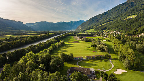 Luftaufnahme des Golfplatzes vor der Kulisse majestätischer Berge unter einem klaren blauen Himmel. Die Szene ist heiter und malerisch. Links im Bild sieht man den Rhein fliessen.