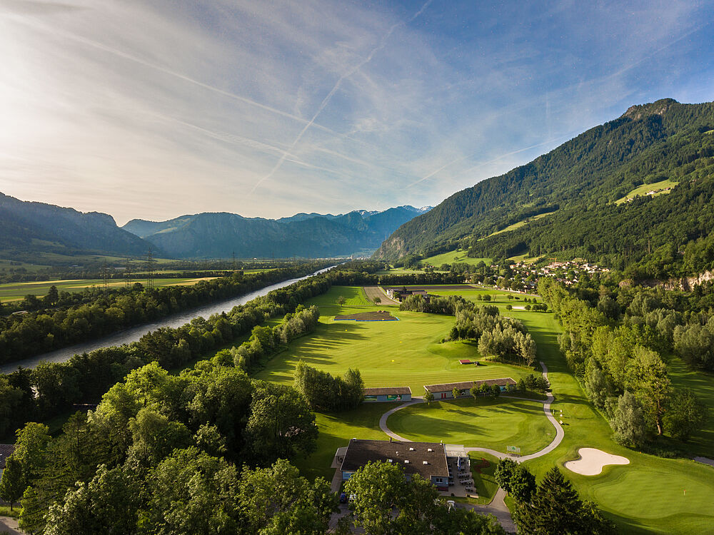 Luftaufnahme des Golfplatzes vor der Kulisse majestätischer Berge unter einem klaren blauen Himmel. Die Szene ist heiter und malerisch. Links im Bild sieht man den Rhein fliessen.