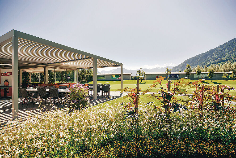 Der Aussenbereich des Golf Bistro mit einer Pergola an einem sonnigen Tag, umgeben von üppigen grünen Rasenflächen und Wildblumen, mit Hügeln in der Ferne unter einem klaren Himmel.