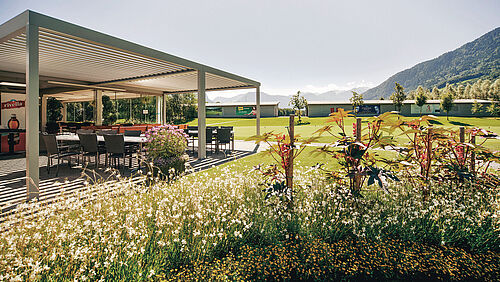 Der Aussenbereich des Golf Bistro mit einer Pergola an einem sonnigen Tag, umgeben von üppigen grünen Rasenflächen und Wildblumen, mit Hügeln in der Ferne unter einem klaren Himmel.