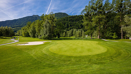 Der Golfplatz mit ordentlich gemähtem Gras, umgeben von Bäumen unter einem klaren blauen Himmel. Berge im Hintergrund, die eine ruhige Atmosphäre schaffen.