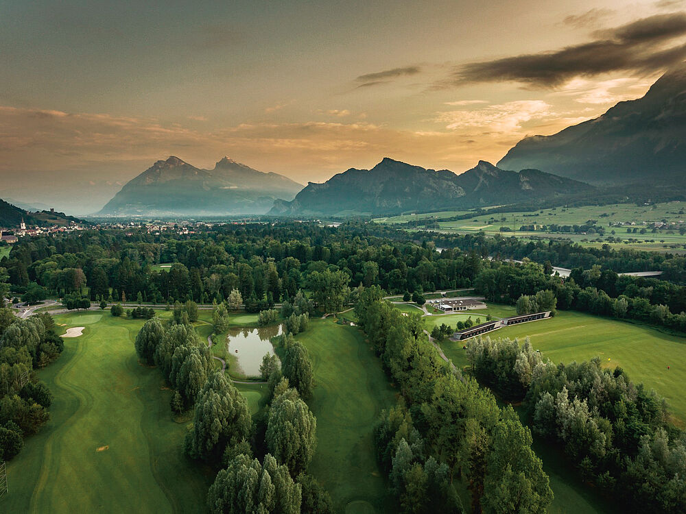 Luftaufnahme des Golfplatzes bei Sonnenuntergang mit baumbestandenen Feldern, fernen Bergen und teilweise bewölktem Himmel, die Gelassenheit und natürliche Schönheit vermitteln.