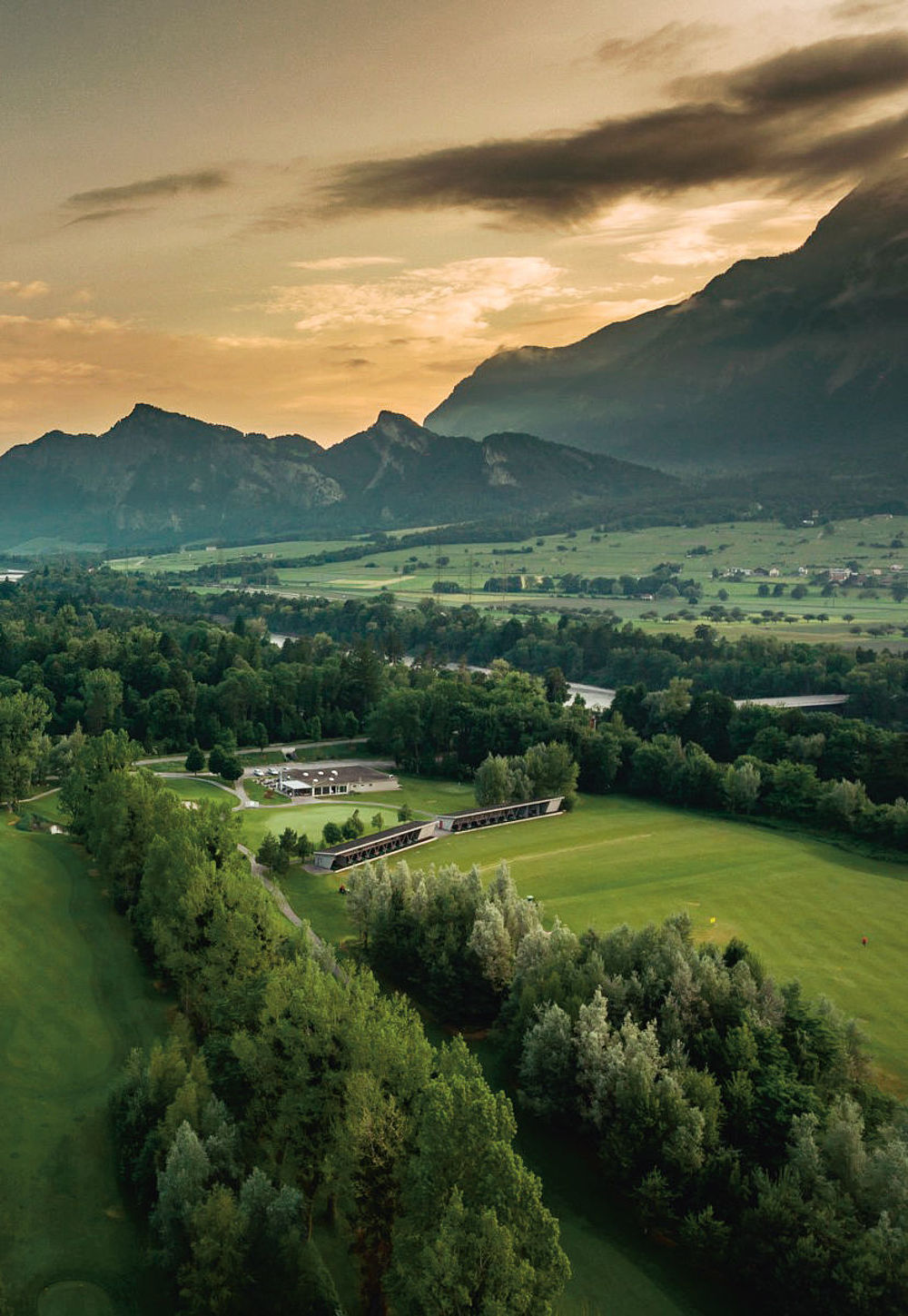 Luftaufnahme des Golfplatzes bei Sonnenuntergang mit baumbestandenen Feldern, fernen Bergen und teilweise bewölktem Himmel, die Gelassenheit und natürliche Schönheit vermitteln.