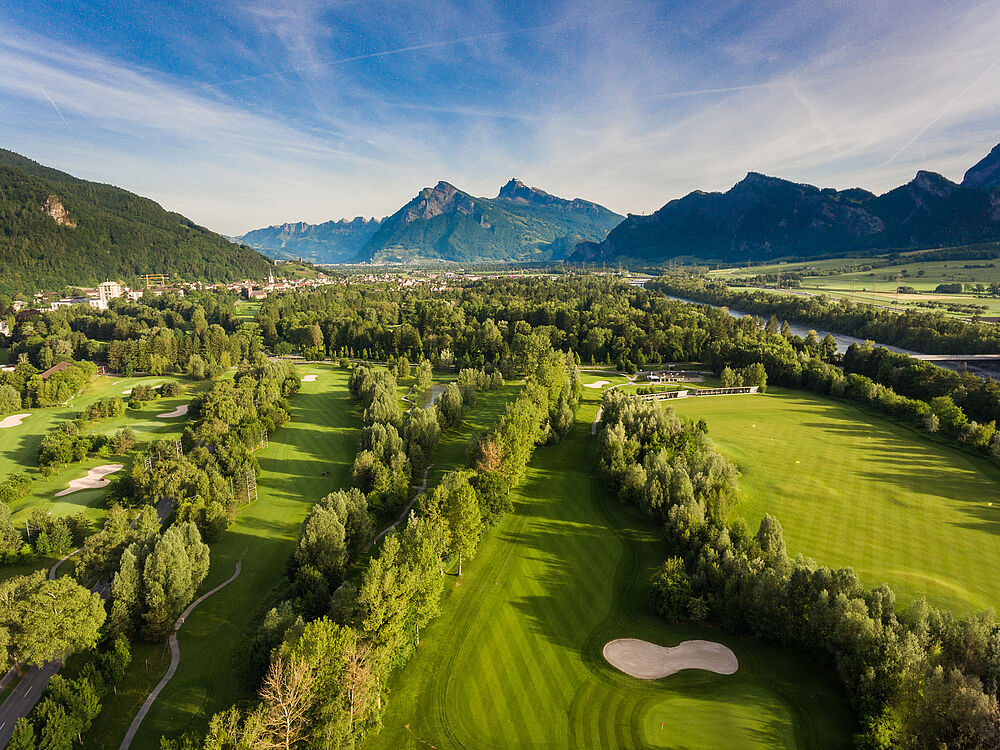 Luftaufnahme des Golfplatzes vor der Kulisse majestätischer Berge unter einem klaren blauen Himmel. Die Szene ist heiter und malerisch.