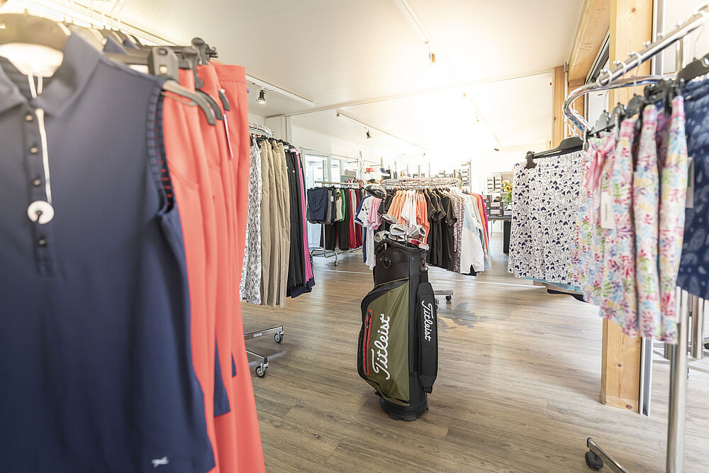 Unser Golf Shop mit Regalen voller farbenfroher Kleidung, darunter marineblaue, korallenrote und gemusterte Artikel. Die helle Beleuchtung schafft eine einladende Atmosphäre.