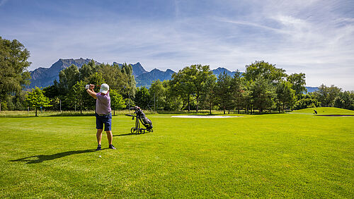Ein Golfer steht auf dem Fairway und holt gerade zum Schlag aus. Hinter ihm steht sein Golfbag und Caddy. Im Hintergrund sind Bäume und die Bergelandschaft zu sehen.